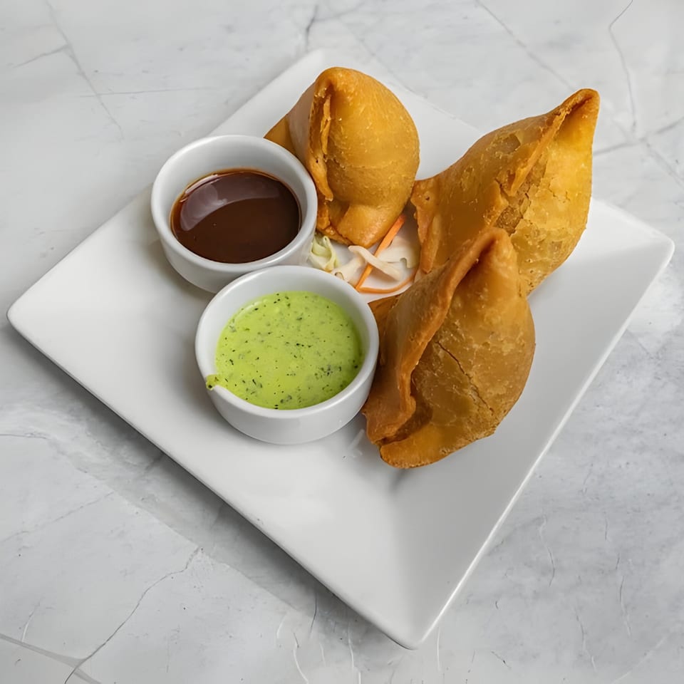 Three golden-brown samosas with two dipping sauces, one dark and one green, on a white plate.