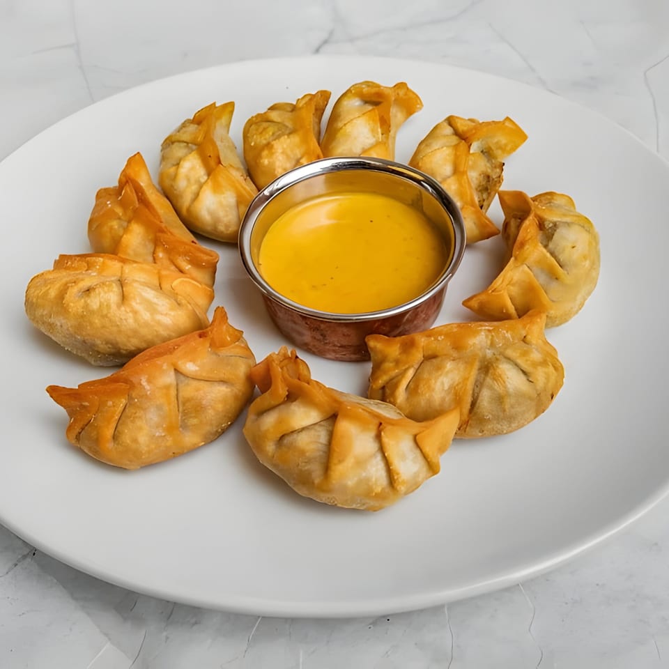 A white plate of golden-brown fried Indian momos arranged around a small metal bowl of yellow dipping sauce.
