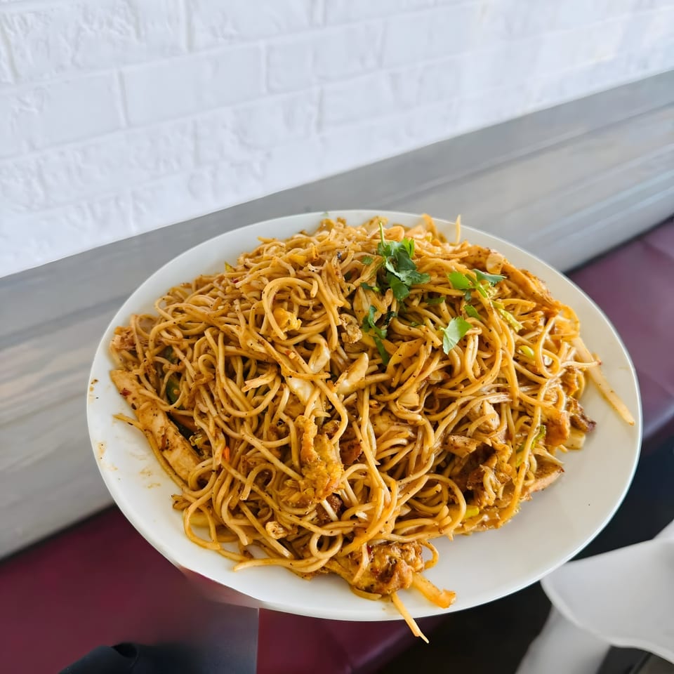 A large plate of Indian-style chicken noodles, garnished with fresh cilantro, against a white brick wall.